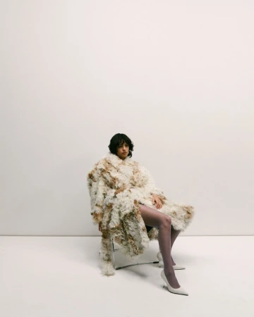 A young adult woman with dark hair sits on a metal chair wearing a fuzzy, oversized coat and matching heels in a brightly lit studio.