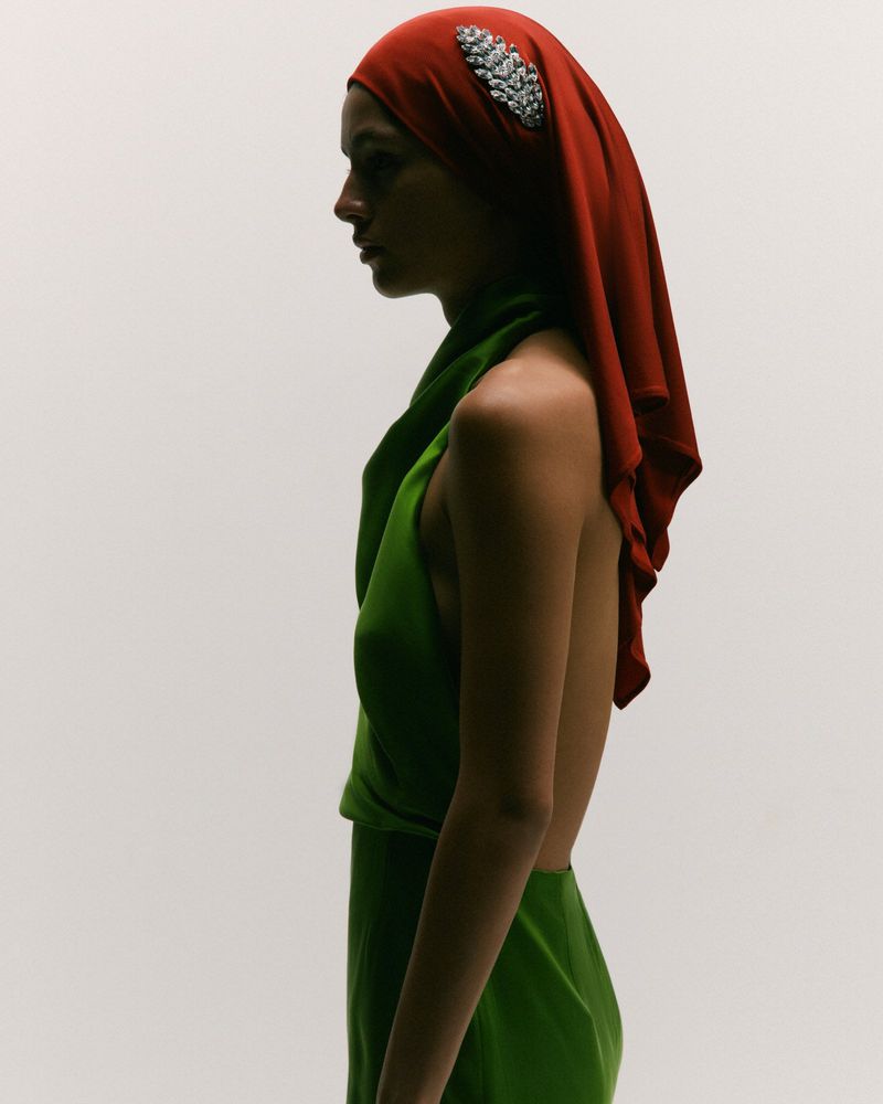 Profile shot of a woman wearing a green gown and a red headscarf with a jeweled ornament against an off-white backdrop.