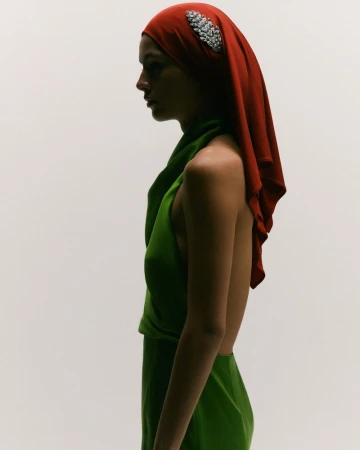 Profile shot of a woman wearing a green gown and a red headscarf with a jeweled ornament against an off-white backdrop.