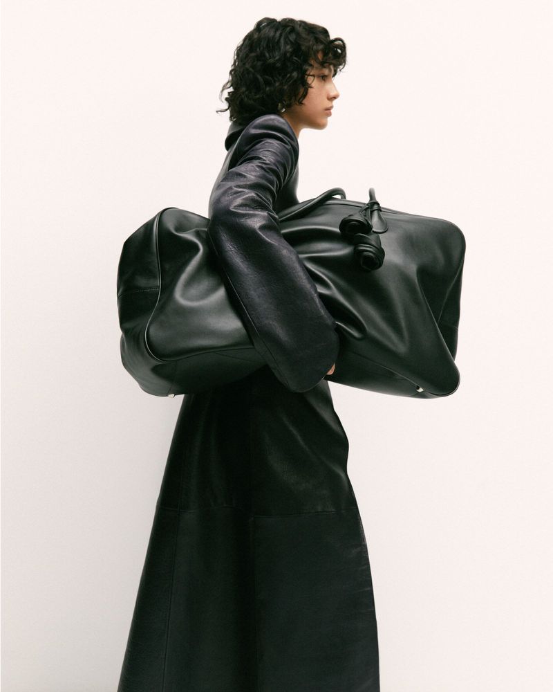 A young adult carries a large black leather duffel bag and wears a matching leather jacket and skirt against a white background.