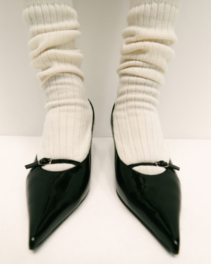 Close-up of two legs with black patent leather pointed-toe heels and leg warmers in front of a white background, fashion style.