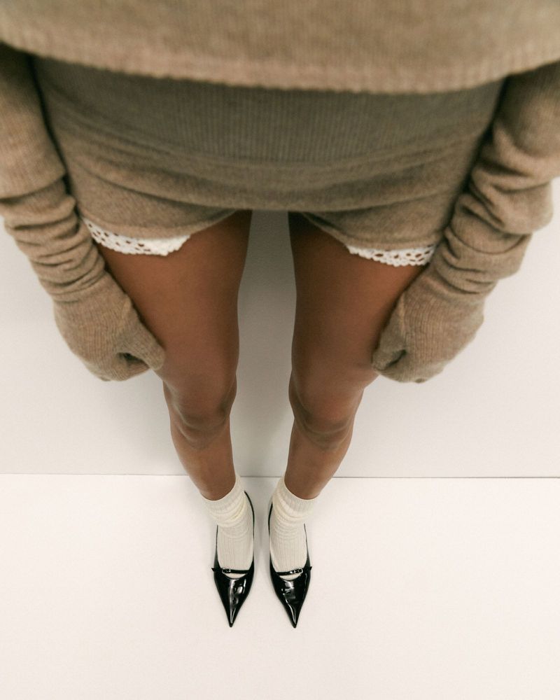 Woman wearing a coordinated tan sweater, skirt and gloves, with ivory socks and black heels, on a white background, shot from above.