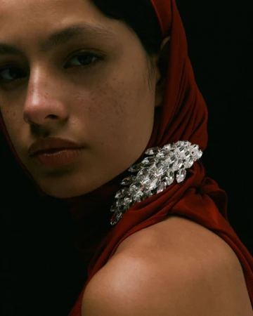 Close-up portrait of a woman wearing a dark red head covering and a large crystal brooch against a black background
