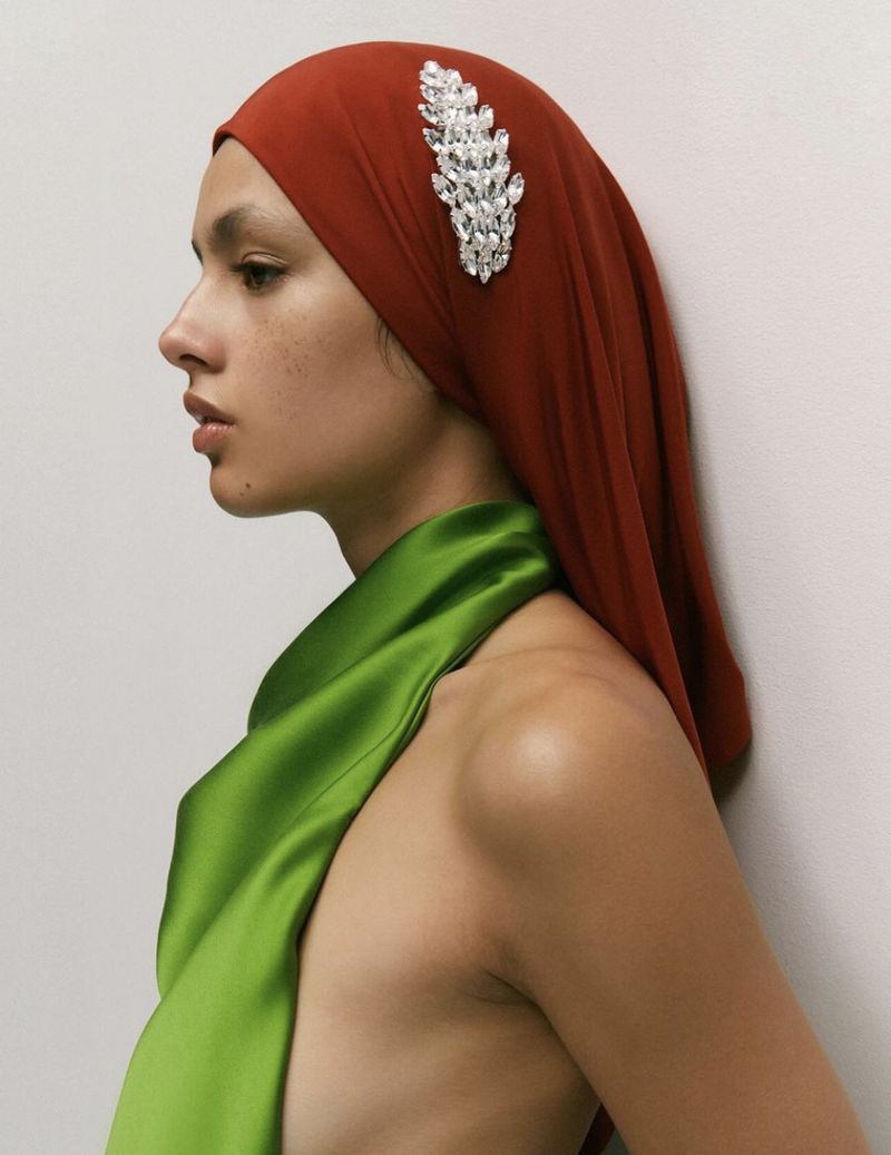 A woman in profile wears a rust-colored headscarf adorned with a jeweled embellishment, and a green satin drape over her shoulder.