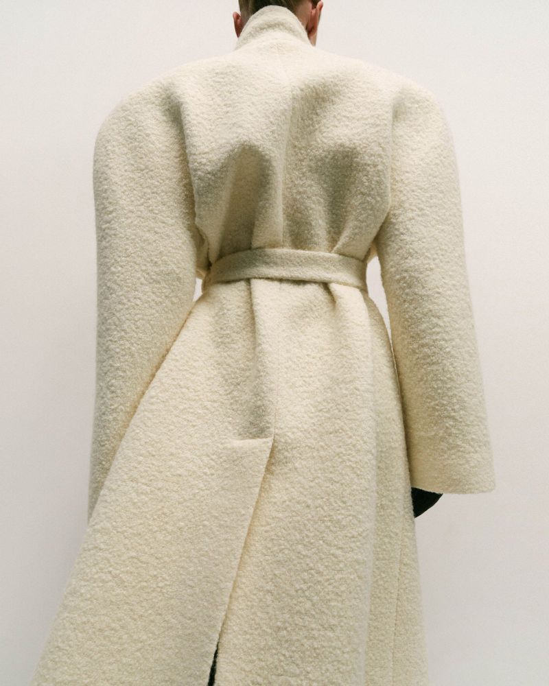 Back view of a slender person in a floor-length, cream-colored bouclé wool coat with a cinched waist against a light neutral backdrop.