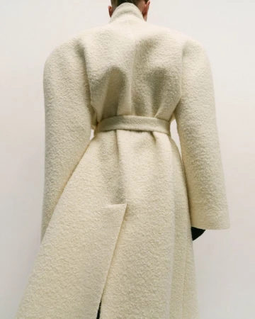 Back view of a slender person in a floor-length, cream-colored bouclé wool coat with a cinched waist against a light neutral backdrop.
