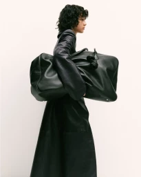 Model with dark curly hair wearing a black leather coat carrying a large black duffel bag against a white background