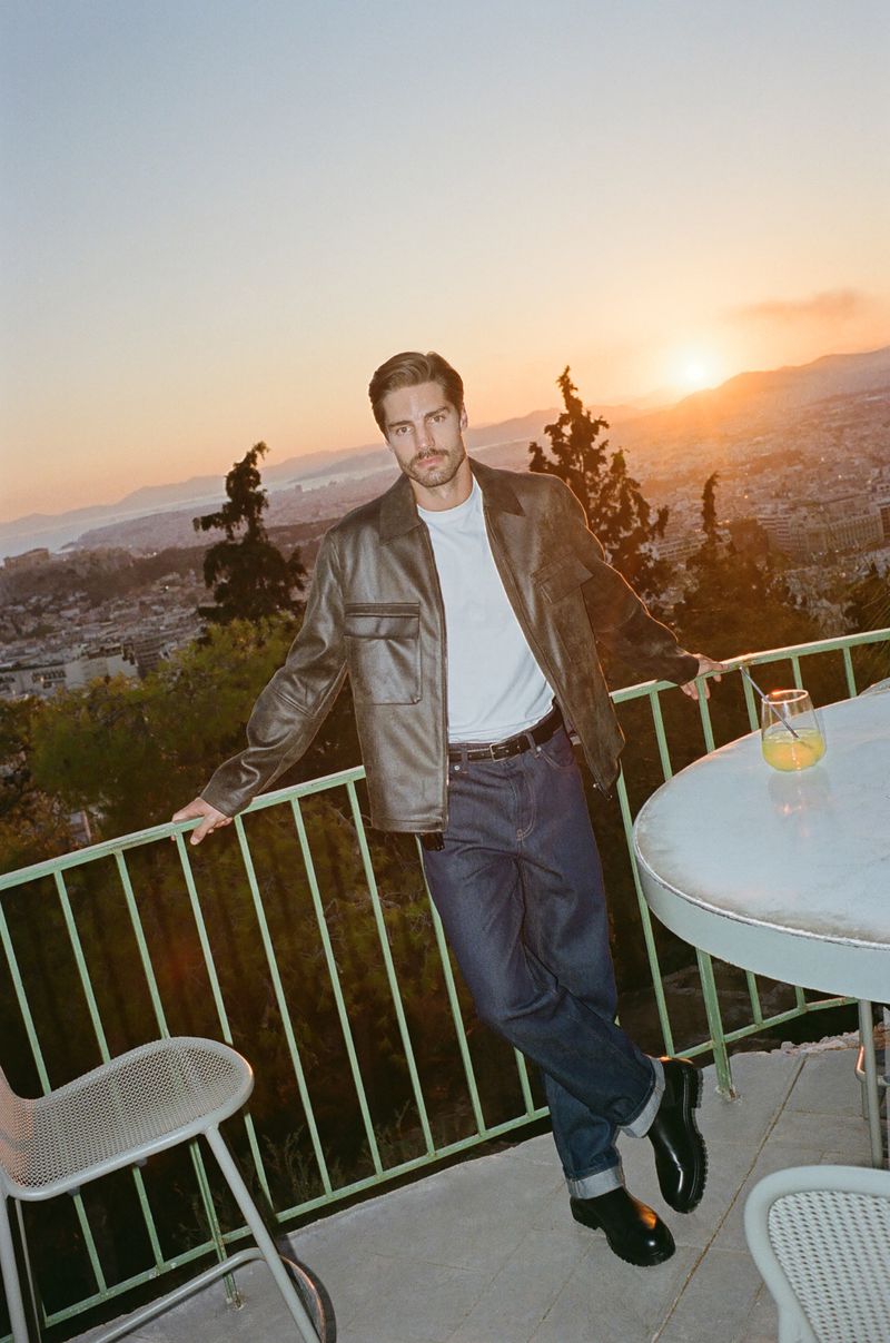 Man in jacket and jeans leans against railing on terrace at sunset. Distant city buildings and mountains are visible.