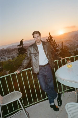 Man in jacket and jeans leans against railing on terrace at sunset. Distant city buildings and mountains are visible.