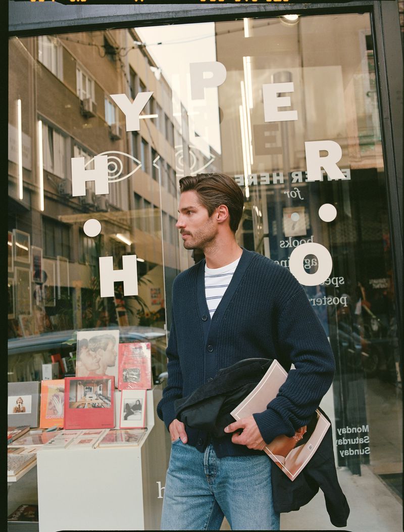 Man stands outside bookshop with books and magazines displayed. He wears a cardigan, jeans, and carries a book with a portrait cover.