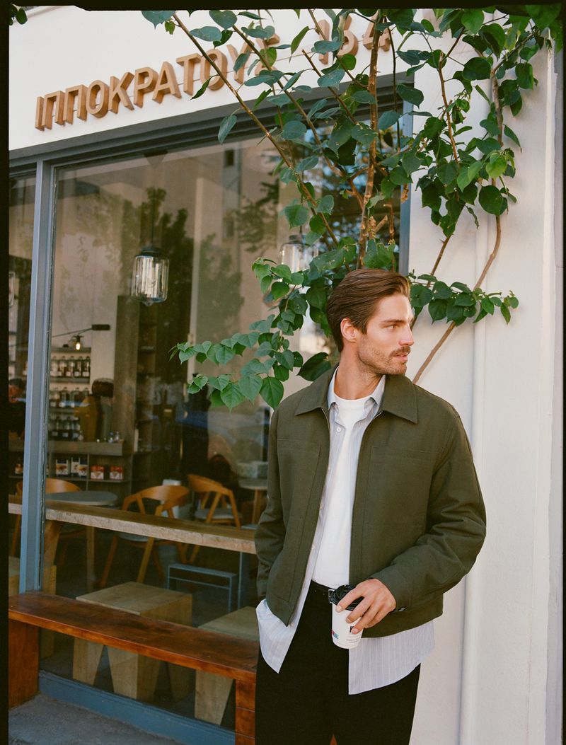 Man in an olive jacket standing outside a cafe, holding a coffee cup, with a tree and the cafe's window in the background.