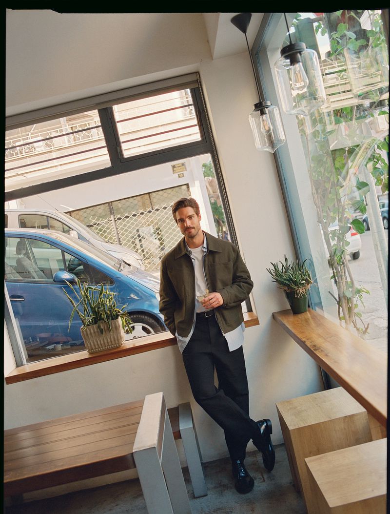 Man in olive jacket, white t-shirt, and black pants poses indoors by window at a cafe, with light streaming in.