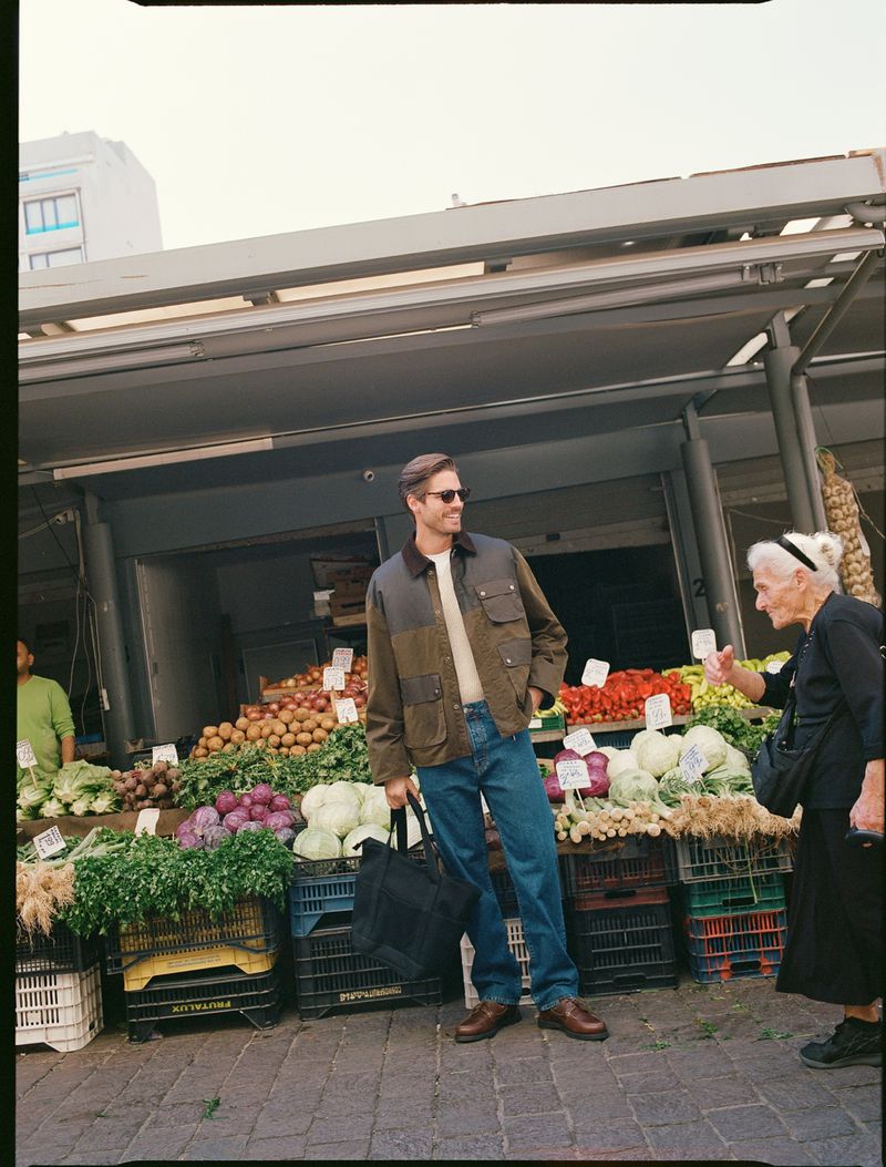 Man in jacket stands near a fruit and vegetable stand with an elderly woman in an outdoor market, under daylight with building in background.