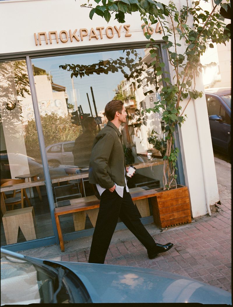 A man walks on a sunny urban street in front of a storefront with reflective glass, wearing casual stylish clothes, on a bright day.