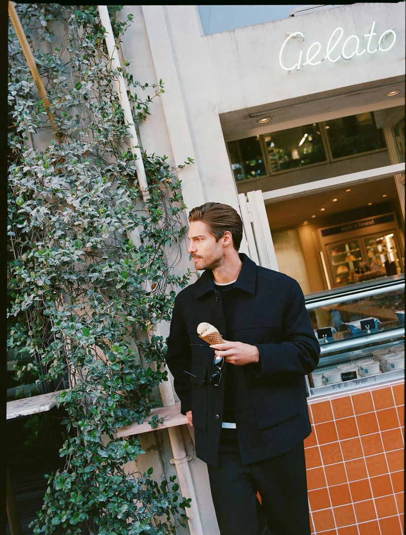 A man in a black jacket and pants stands outside a building with a Gelato sign, holding an ice cream cone and eyeglasses. The building has green vines.