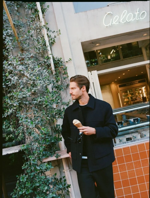 A man in a black jacket and pants stands outside a building with a Gelato sign, holding an ice cream cone and eyeglasses. The building has green vines.