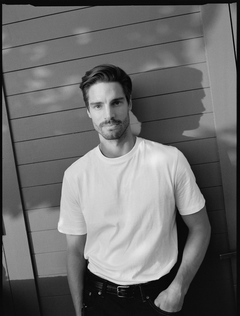 Monochrome portrait of adult male in white t-shirt and black jeans, hand in pocket, standing against a slatted wall casting a shadow.