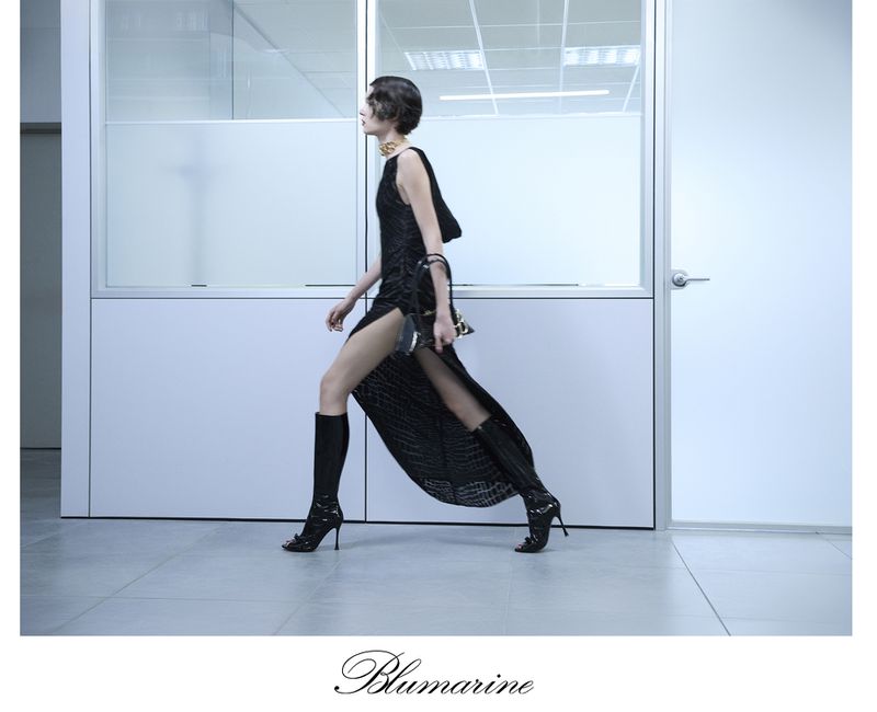 A woman in a black dress and boots walks in a modern, minimalist hallway, carrying a purse. The image is cool-toned and fashion-forward.