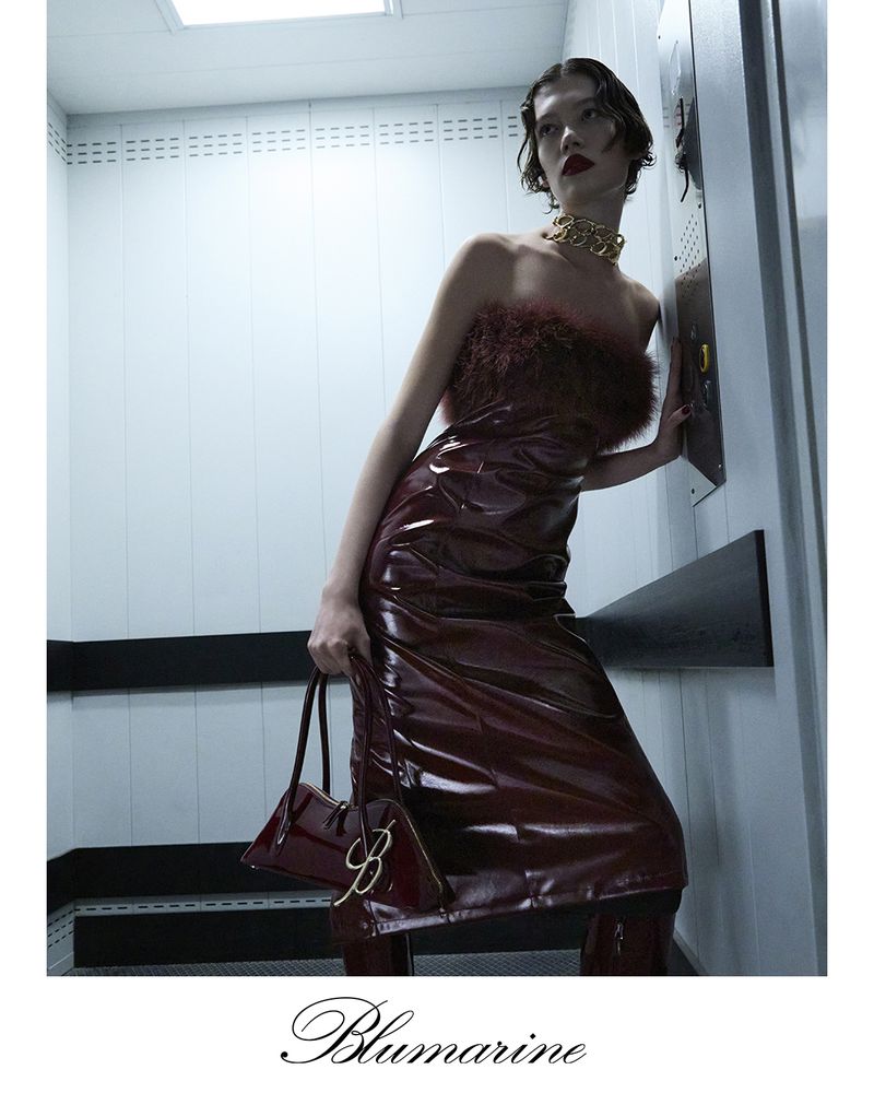 A slender woman in a burgundy latex dress and gold necklace holds a purse while standing in a neutral-toned elevator.