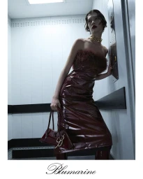 Model in a glossy burgundy feather-trimmed dress holding a matching handbag inside a white elevator interior