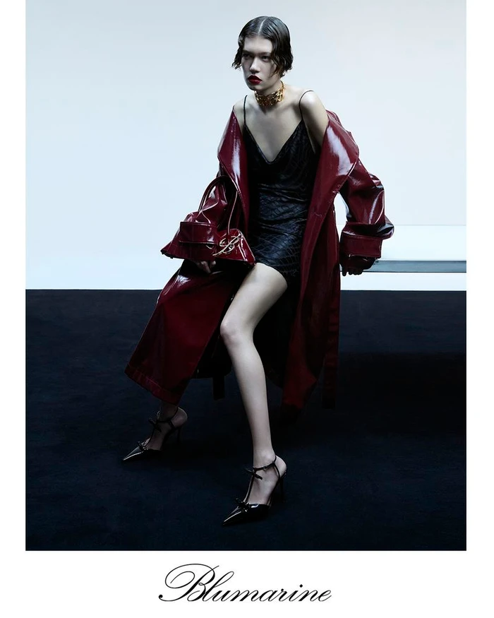 Model in shiny burgundy coat and black dress holding handbag seated in studio with white backdrop and black floor