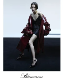 Model in shiny burgundy coat and black dress holding handbag seated in studio with white backdrop and black floor
