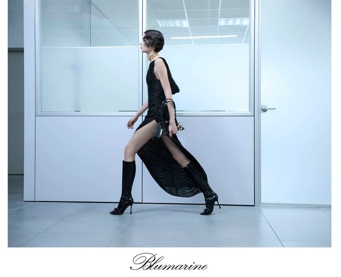 Model wearing a black dress and boots walking past a white glass office partition for Blumarine Pre-Fall 2024 collection.