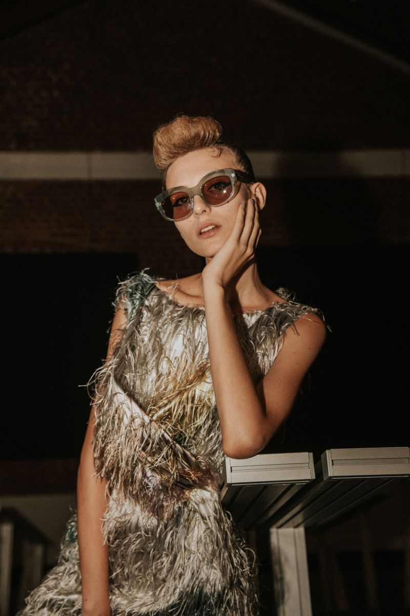 Woman in a feather dress and stylish sunglasses poses indoors, hand to face, with soft, diffused lighting highlighting fashion and style.