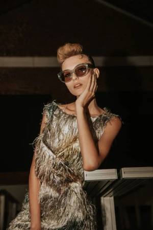 Woman in a feather dress and stylish sunglasses poses indoors, hand to face, with soft, diffused lighting highlighting fashion and style.