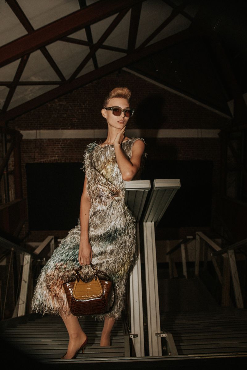 Model in textured fringe dress wearing sunglasses holding a brown handbag on a staircase backstage at Antonio Marras SS25.