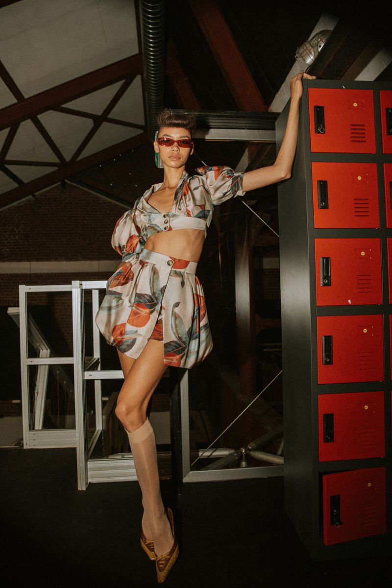 Woman poses in a patterned two-piece outfit, sheer socks, and heels in a room with lockers, showcasing a blend of fashion and edgy style.