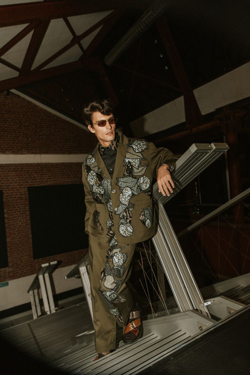 Man in olive green patterned suit and sunglasses poses on metal structure in an industrial indoor space with brick wall backdrop.