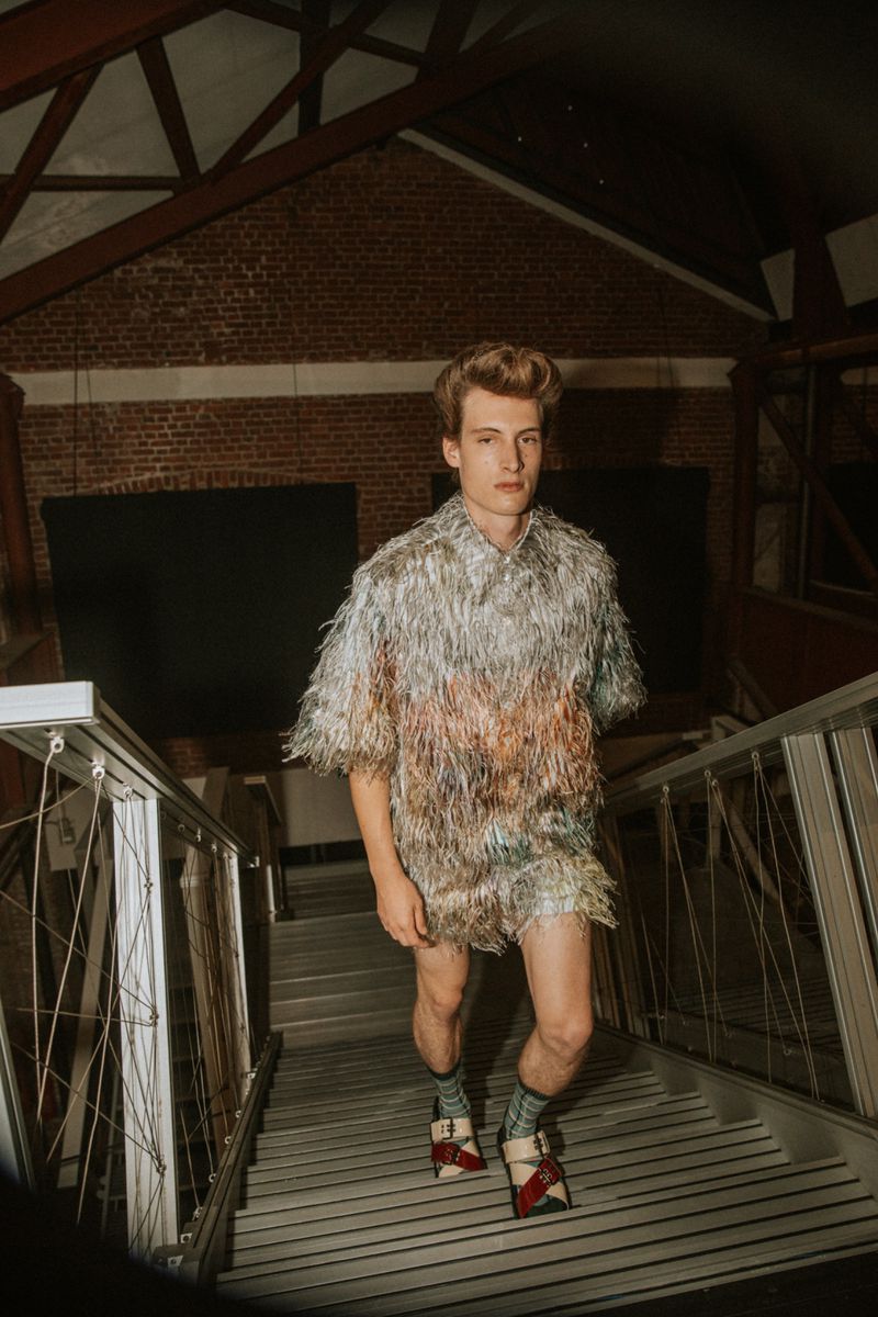 Male fashion model poses indoors on steps wearing shimmering silver fringed top, matching shorts, socks, and red-and-white buckled shoes.