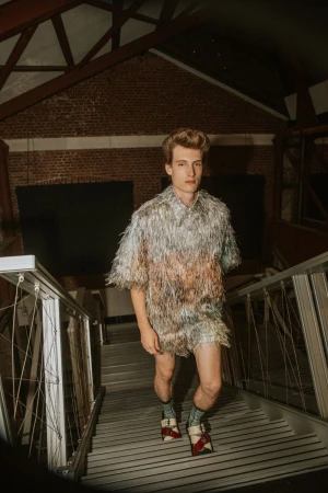 Male fashion model poses indoors on steps wearing shimmering silver fringed top, matching shorts, socks, and red-and-white buckled shoes.