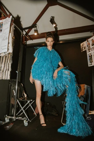 A runway model in a blue fringe dress poses in a backstage setting with racks of clothes and lighting equipment in the background.