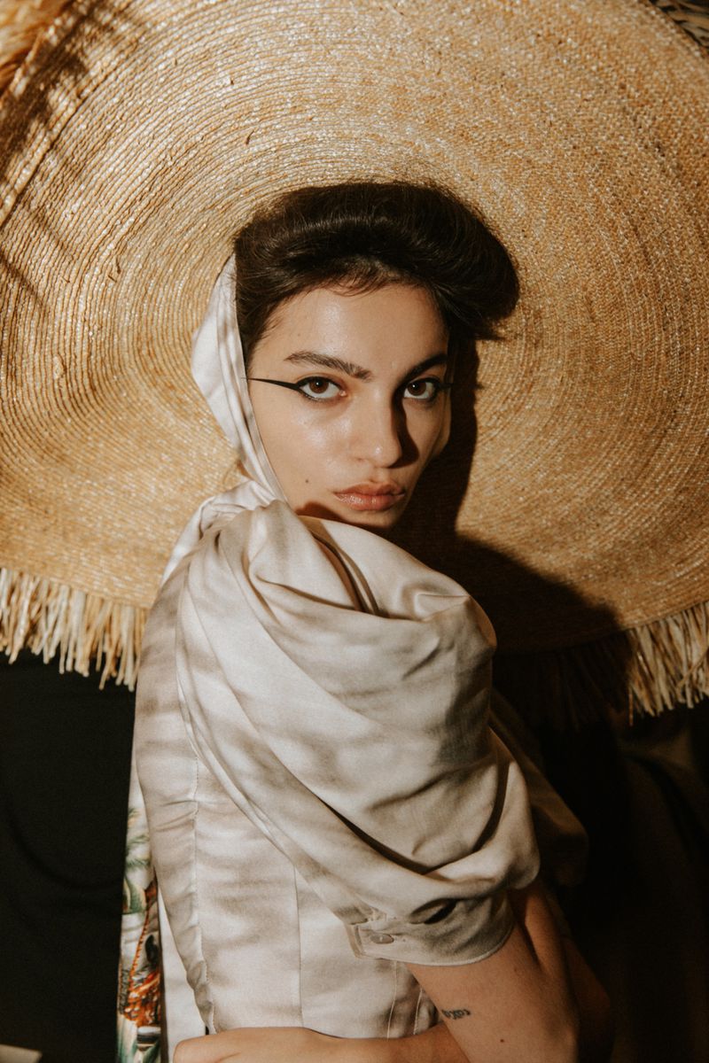Woman in straw hat and headscarf gazes at camera. Close-up fashion headshot with neutral tones and soft lighting in an indoor setting.