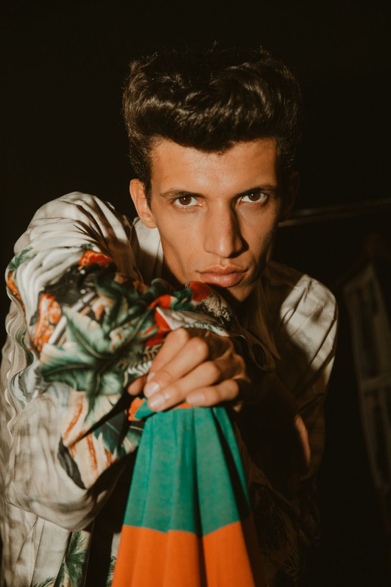Portrait of a young adult male with dark hair, wearing a floral shirt and holding a colourful umbrella against a dark background.