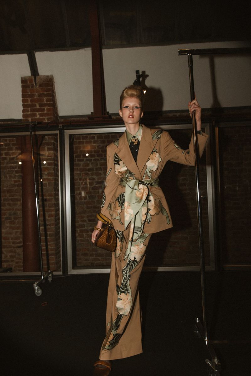A stylish woman in a floral suit poses indoors, holding a metallic rack, with a brick wall visible in the background, showcasing fashion.