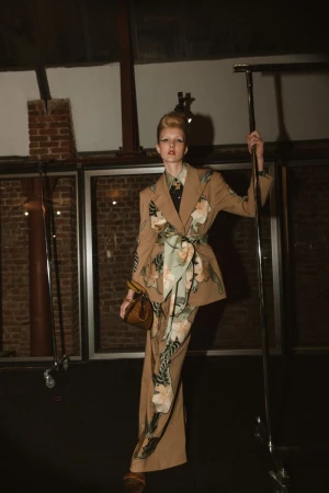 Model in a patterned floral tan suit holding a handbag inside a backstage room with exposed brick walls