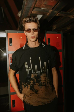 Model wearing dark tinted sunglasses and a black graphic t-shirt posing against red lockers backstage at Antonio Marras SS25