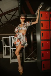 Model leaning against a stack of red lockers wearing a patterned cropped top and skirt set with sheer knee-high socks.