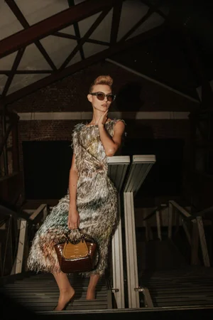 Model in textured fringe dress wearing sunglasses holding a brown handbag on a staircase backstage at Antonio Marras SS25.