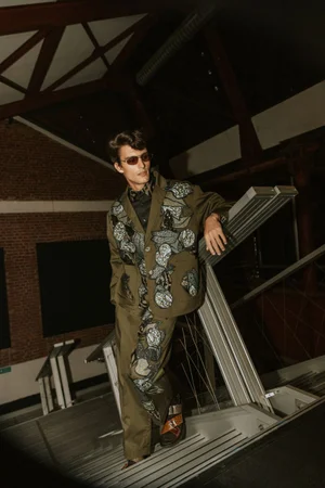 Model wearing an olive green patterned suit and sunglasses leaning on metal stairs indoors.