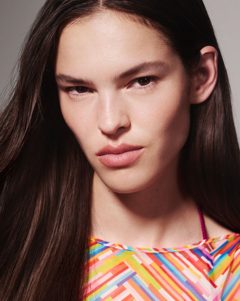 Close-up studio portrait of a woman with long brown hair wearing a multicolored geometric-patterned top against a grey background.