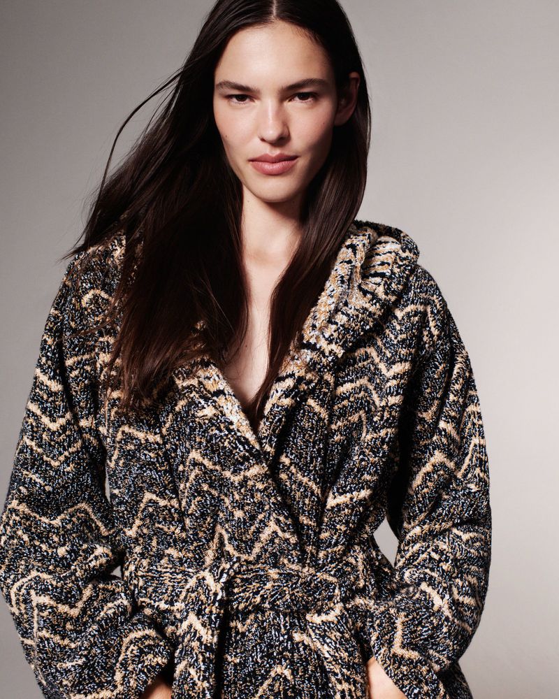 Brunette adult woman poses in a hooded robe with a black, brown and beige geometric pattern in a studio setting.