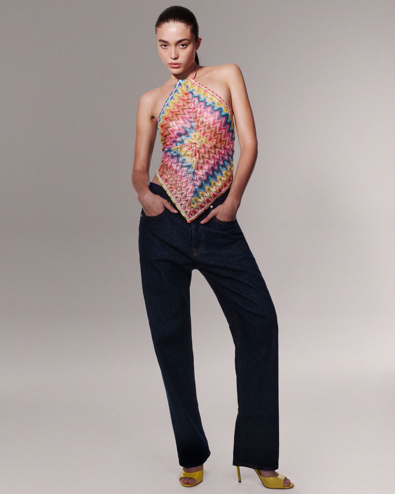A stylish young woman in a colorful top and dark jeans stands in a studio setting, striking a pose against a neutral backdrop.