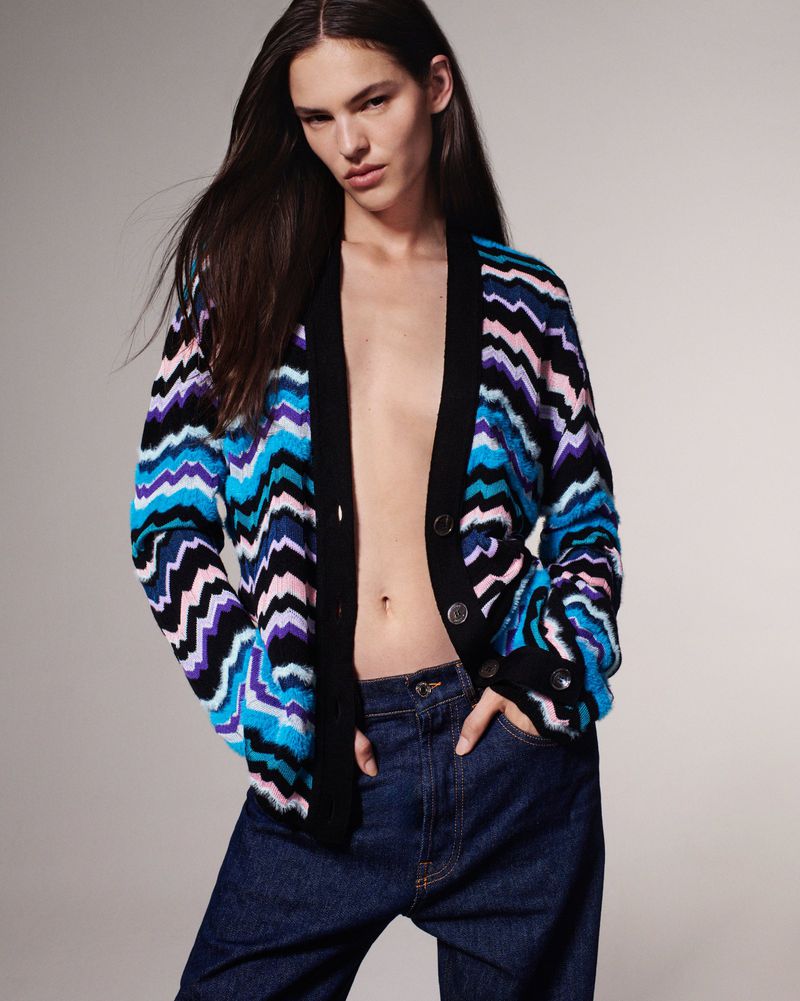 Brunette model wears an open, colorful chevron-patterned cardigan with dark-wash jeans in a studio shot against a pale grey background.