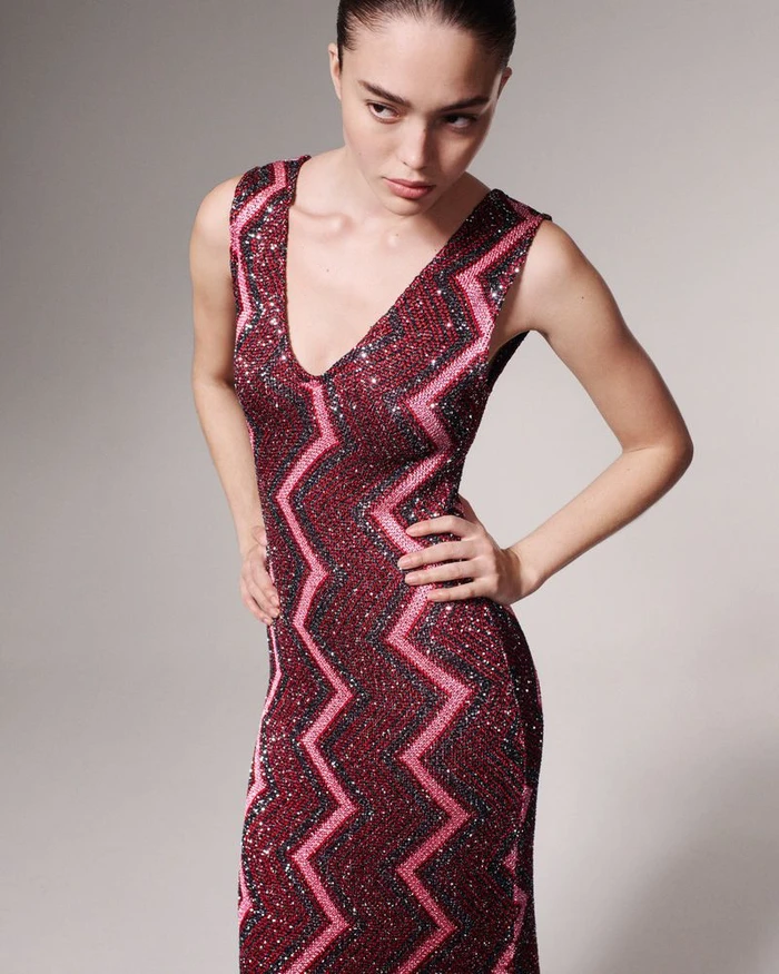 Model wearing a sequined pink and burgundy chevron dress standing against a plain neutral backdrop