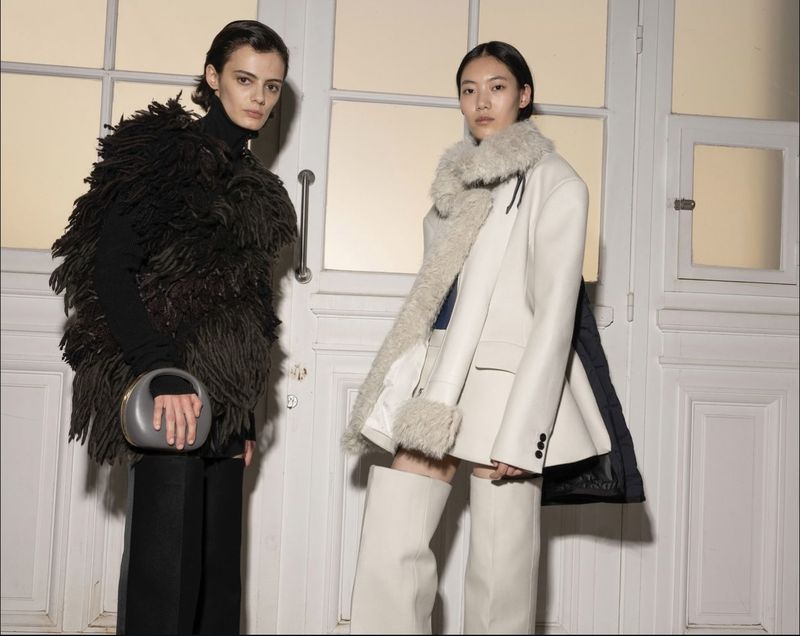 Two women in fashionable outfits in a photo studio. One wears a feathery black top and the other wears a white blazer with fur collar.