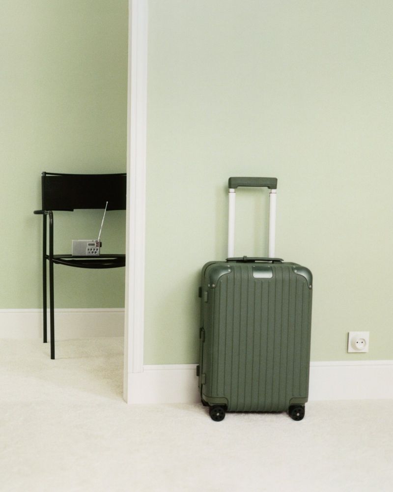 Green rolling suitcase against light green wall in minimalist interior with a chair and radio visible through a doorway. Interior design.